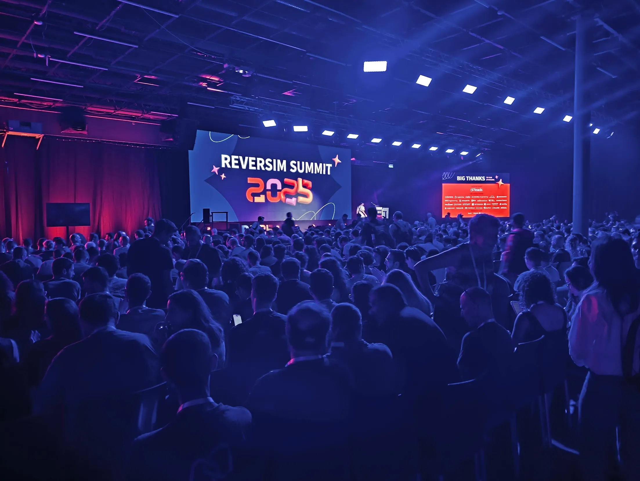 Packed main stage audience under stage lighting with the sponsor thank-you wall to the right